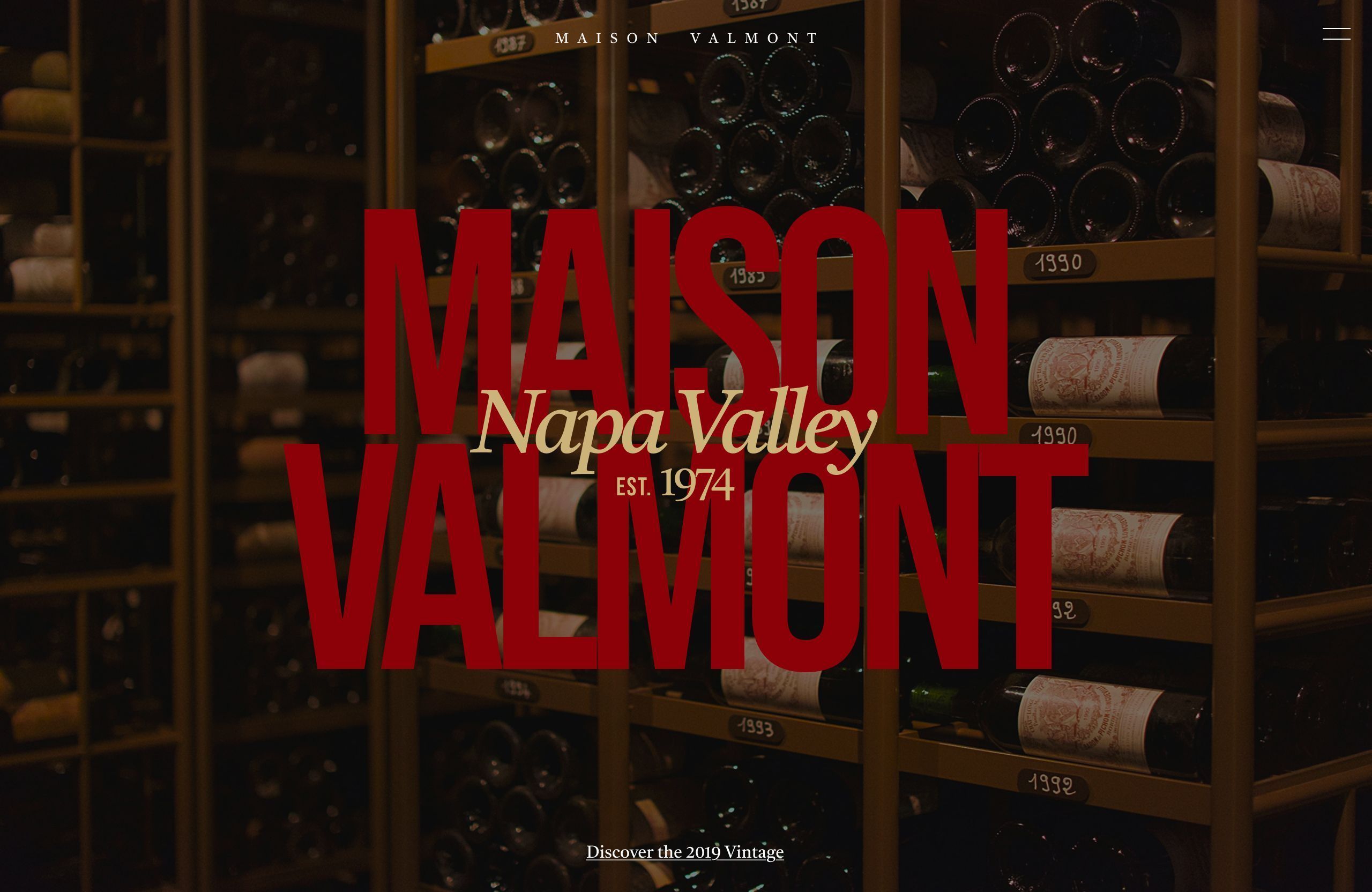 Dark wine-cellar hero with racked vintage bottles; oversized red MAISON VALMONT wordmark overlaid with a cream Napa Valley script and EST. 1974.